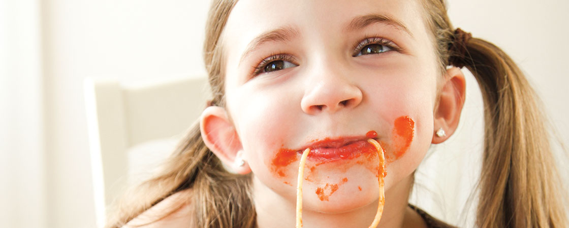 A smiling girl with piece of spaghetti hanging from her mouth and sauce on her cheeks